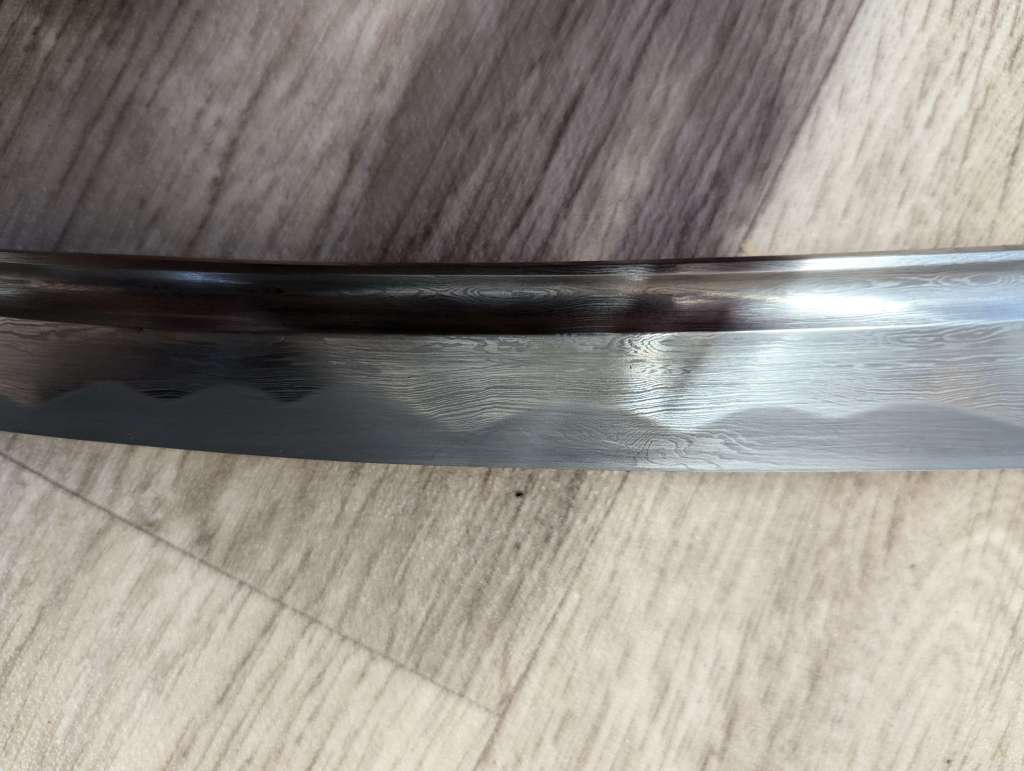 Close up of a katana blade. The hamon has an irregular pattern of curves, somewhat resembling a mountain range.